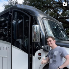 A student in grey shirt giving a thumbs-up in front of a black and white bus
